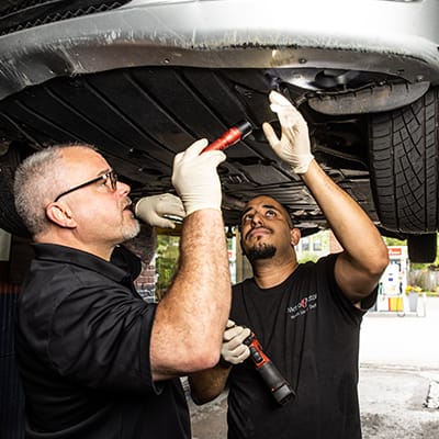 Two auto repair technicians inspect the underside of a vehicle in Alexandria
