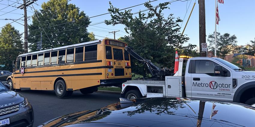 A Metro Motor tow truck tows a school bus on a busy street.